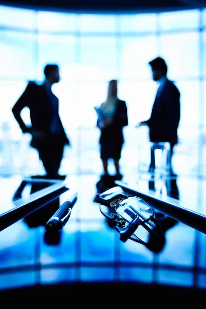 Três executivos conversando de pé em uma sala de vidro.